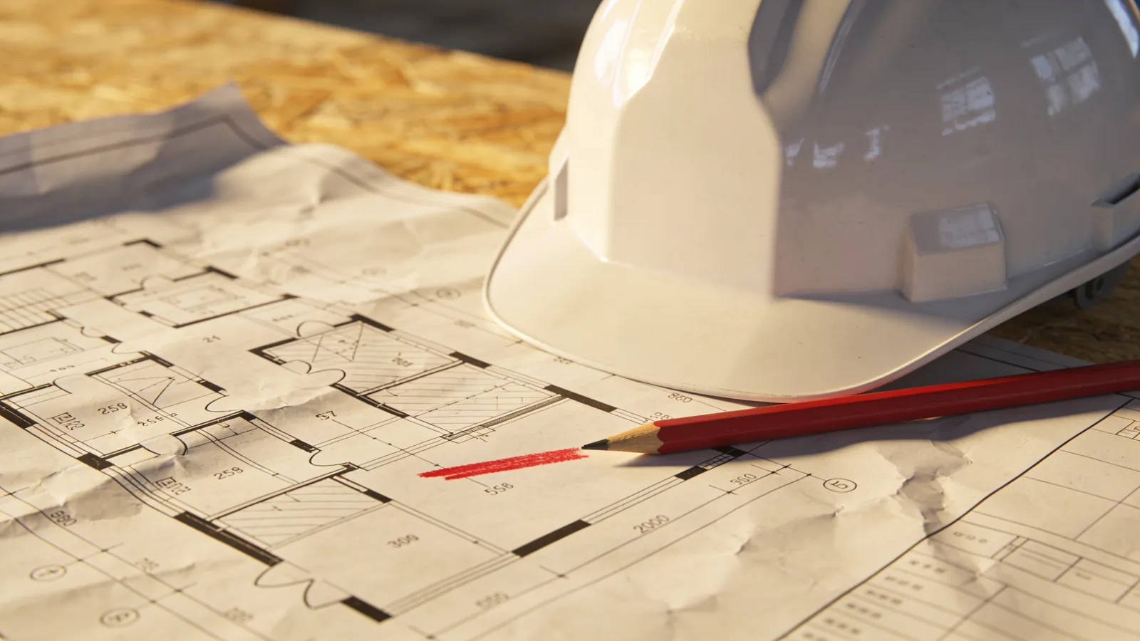 Construction floor plans on a site table with a red pencil marking a change and a white hard hat beside them