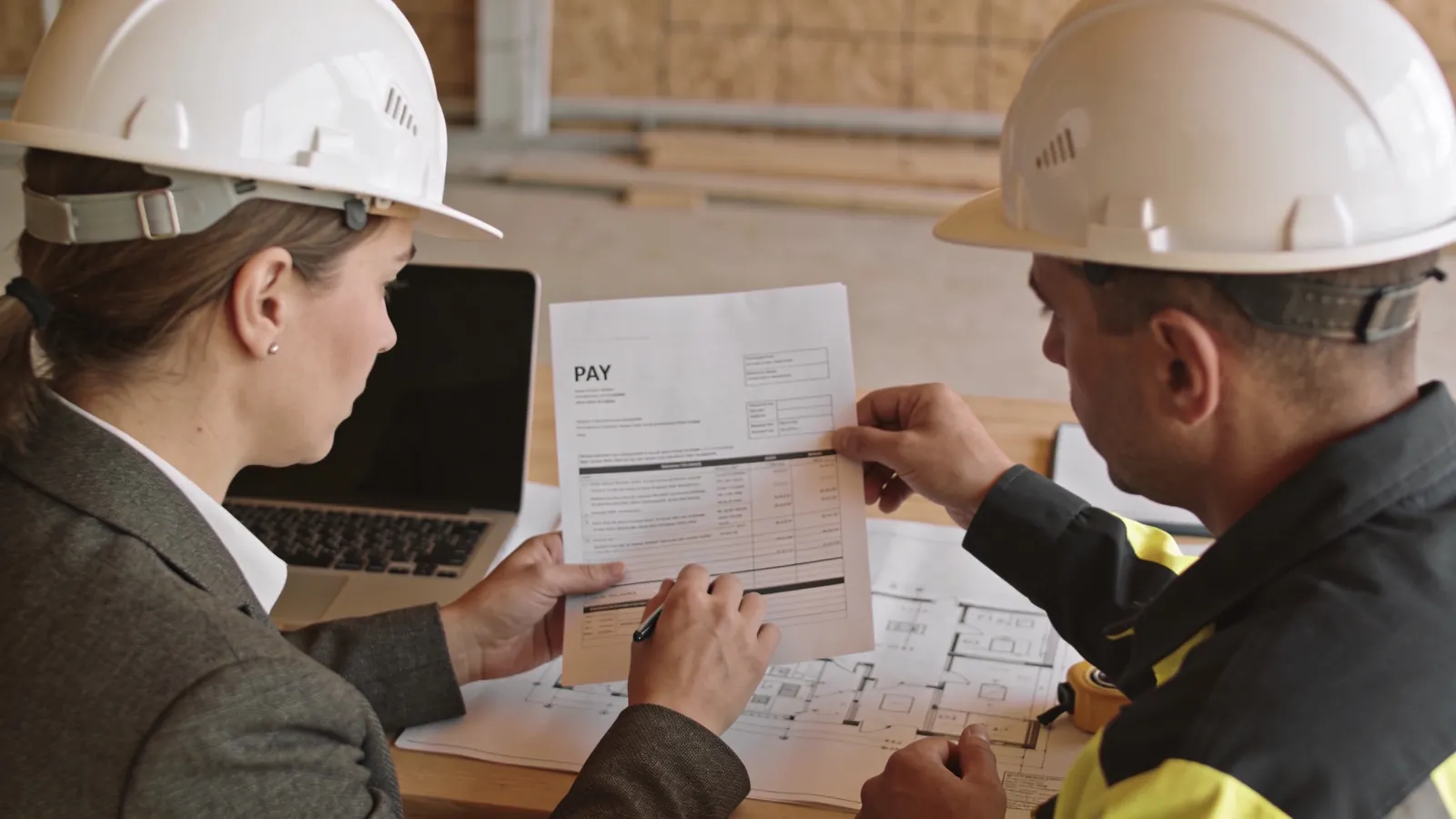 An owner's representative in a blazer and a white hard hat reviewing a printed pay application with a contractor's project manager at a site office table, with floor plans and a laptop in front of them